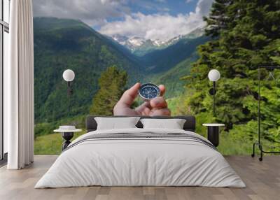 a man holds a compass in his hand against the backdrop of nature, a green forest and mountains in fluffy clouds Wall mural