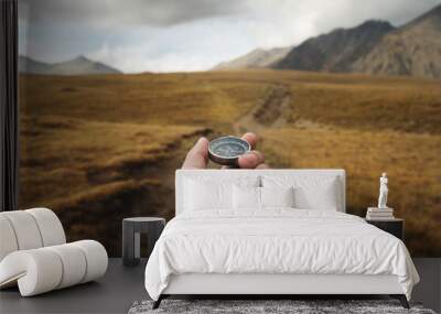 Man's hand holding a magnetic compass first-person view against the background of a high-altitude path and mountains Wall mural