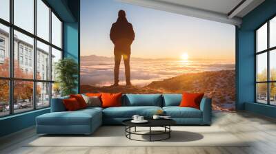 View From the back. A lonely standing man high in the mountains looks at the setting sun and the sunset horizon with a valley filled with clouds. The concept of tourism travel and male loneliness Wall mural