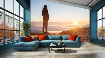 View From the back. A lonely standing man high in the mountains looks at the setting sun and the sunset horizon with a valley filled with clouds. The concept of tourism travel and male loneliness Wall mural