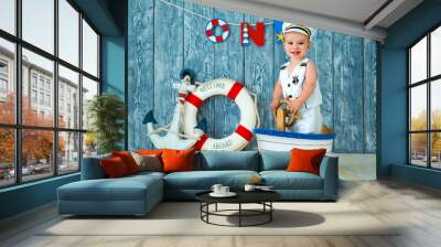 Photoshoot for a boy of one year. Little sea captain, sailor on toy ship with steering wheel. Sea anchor and lifebuoy on gray wooden background Wall mural