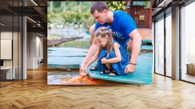 hungry gold asian fish eats food from bottle in the pond. little beautiful girl with Dad feeds fish Wall mural