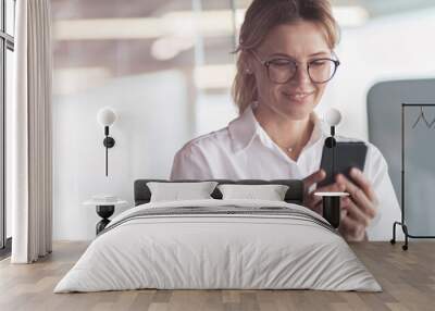 Businesswoman wearing glasses with mobile phone in hand standing in office. Blurred background Wall mural