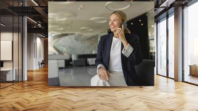 Cheerful businesswoman talking phone sitting on modern office background and looks away Wall mural