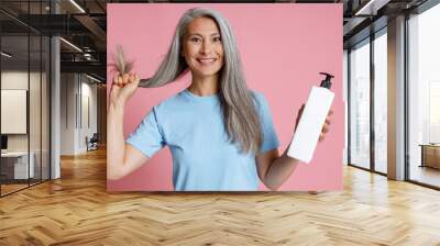 Joyful middle aged Asian woman shows healthy silver hair and blank dispenser bottle of cosmetic products on pink background in studio. Mature beauty lifestyle Wall mural