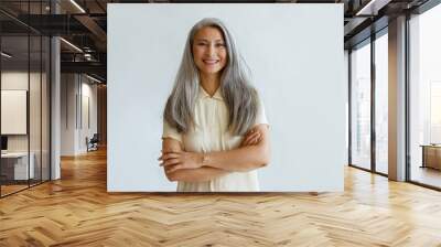 Portrait of smiling Asian woman with long hoary hair and crossed arms standing on light grey background in studio. Mature beauty lifestyle Wall mural