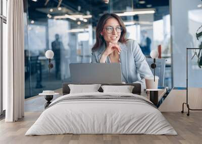 Smiling businesswoman with glasses working on laptop and looking out the window at her workplace in modern office. Blurred background. Wall mural