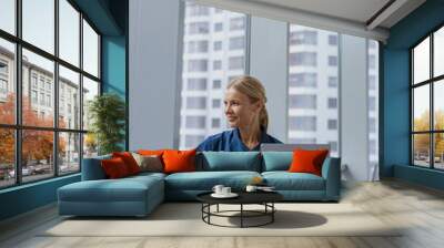 Smiling female healthcare worker using laptop while working at doctor's office and looking window Wall mural