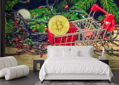 Bitcoin in a mini shopping cart on the background of Christmas tree branches with lights, next to small gifts. The concept of Christmas, concept of purchasing cryptocurrency Wall mural