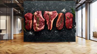 A variety of raw beef steaks for grilling with seasonings: T-bone, striploin steak, ribeye. Top view. On a black stone background. Wall mural