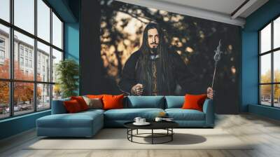 A man with long hair and beard in a black cloak in the image of a sorcerer warlock on Halloween. Dark magician the man in the background of the forest. A wizard in a dark forest with a spear Wall mural