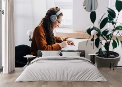 Busy young woman university student wearing headphones using laptop e-learning, writing notes, studying online education seminar class by webinar, learning online seminar sitting at the home table. Wall mural