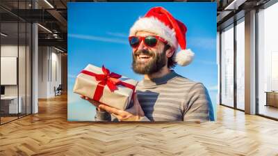 Festive man with Santa hat and sunglasses holds a Christmas gift against a bright blue sky, embodying holiday cheer and warmth. Wall mural