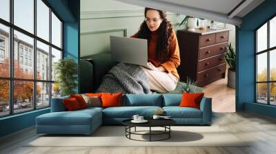 Relaxed young woman sitting on sofa holding mobile phone using cellphone and laptop technology doing online ecommerce shopping, texting messages relaxing on couch in cozy living room home interior. Wall mural