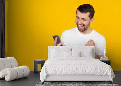 Young man with a beard in a white t-shirt shouts with pride and celebrates victory and success with a smartphone, very excited, jubilant emotion. Stands on isolated yellow background Wall mural