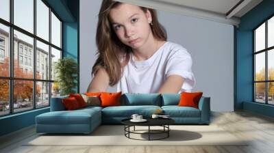 A young girl in casual attire looks thoughtfully into the camera, seated against a neutral background. Youth introspection reflects the complexities of adolescence in modern society. Wall mural