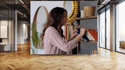 young modern woman in knitted dress chooses a book at home library Wall mural