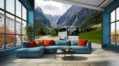 Blue ice tongue of Jostedal glacier melts from the giant rocky mountains into the green valley with waterfalls. Big white tourist bus rides on the road in Norway in summer cloudy day Wall mural