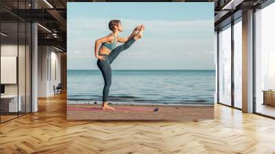 a young attractive woman, of Asian appearance, practicing yoga, performs a stretching exercise, on the beach. pose with an outstretched leg Wall mural