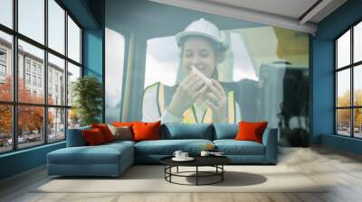 smile woman foreman manager in reflective vest safety jacket sits down in large forklift for relaxing eats bread snacks during brunch break, Foreman have lunch in Forklift Wall mural