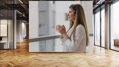 Young woman thinking and looking away with cup of tea or coffee with blurred city background. Girl dressed in white shirt with tea at balcony. Breathing fresh in the morning. Relax comfort concept Wall mural