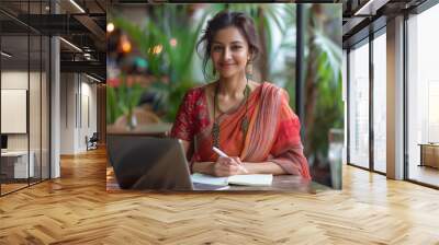 An Indian woman in a saree writing on a notepad at a table with a laptop and water Wall mural