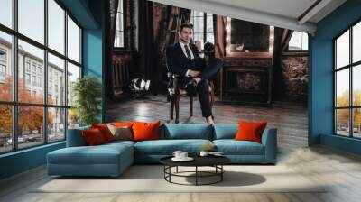 Fashion luxury man sitting on chair. Stylish businessman in full suit posing in interior room. Young handsome male model. Business concept. Success lifestyle Wall mural