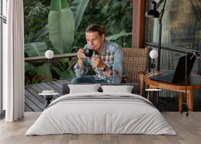 Handsome young man relaxing and working on laptop computer at home balcony with tropical nature, drink coffee Wall mural