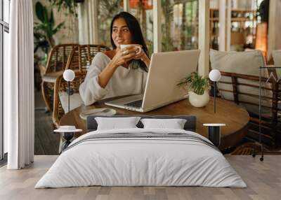 Image of happy brunette  woman using laptop while sitting at cafe with cup of coffee. Young caucasian  woman sitting in a coffee shop and working on laptop. Wall mural