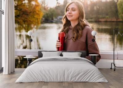 Young woman in hoodie holding a red thermo mug in an autumn park. Sunny weather. Fall season. Wall mural