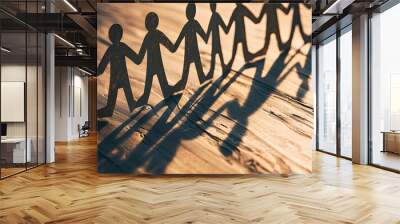 Paper cut-out of people holding hands in a chain, casting long shadows on a wooden surface, with a blurred background to emphasize unity and connection within a community or group. Wall mural