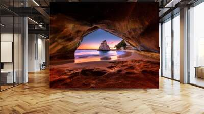 A scenic vista of a beach framed by a natural stone archway at sunset, revealing a rocky island and the vast ocean beyond. Wall mural