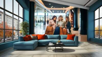 Three women are standing in a mall, looking at shopping bags. They are happy and excited about their purchases. Wall mural