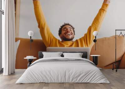 A happy man with a laptop is sitting on the sofa in the living room at home, stretching his arms up to the side and smiling while working online from the house using a computer. Wall mural