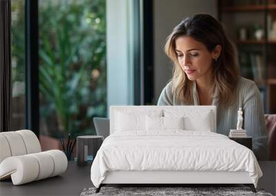 A woman sitting at her desk in front of an open laptop, working on marketing for a small business. In the background is a modern interior design office with a glass door leading to an outdoor area. Wall mural