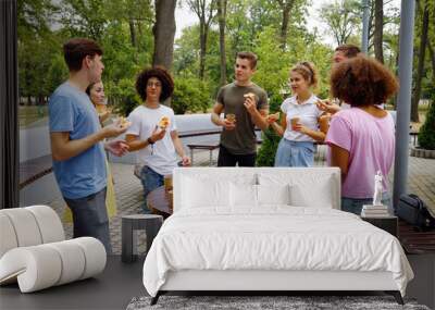 Group of diverse young people in casual wear gathering around table in park and eating pizza, bags and exercise book lying beside. Arc shot students having snack together after classes Wall mural