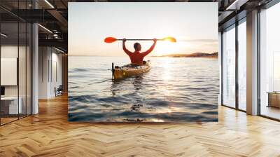 Rear view of man holds kayak paddles high. Active senior in a kayak on the sunset sea  Wall mural