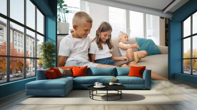 Three young siblings on the couch, two using a tablet computer 
 Wall mural