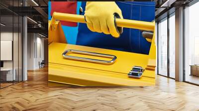 A worker wearing gloves holds a yellow tool box in one hand, representing readiness and safety for a construction or repair task, emphasizing protection and efficiency (6) Wall mural