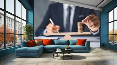 businessperson stands blurred in the background while focusing on balancing stacked coins on a seesaw, symbolizing financial stability, balance, and economic decision-making in business Wall mural