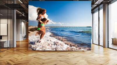 Young blond woman in white bikini and small happy girl entering sea and having fun with sandy beach at background on clear summer day. Travelling, vacations, relaxation, family weekend concept Wall mural