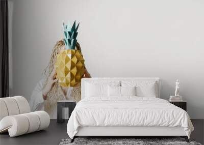 Woman holding paper pineapple in front of face, light grey background, no face trend concept. Young Female with long curly hair dreadlocks hiding behind pineapple. Creative summer concept Wall mural