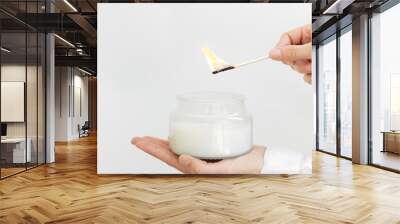 Womans hand uses match to light aromatic candle in glass jar with natural ingredients on white background. Wall mural