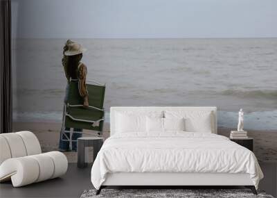 A woman is standing on the beach holding a green beach chair. The beach is calm and peaceful, with the ocean in the background. The woman is enjoying the serene atmosphere Wall mural