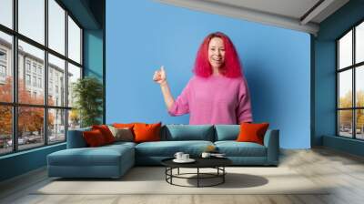 Woman with pink hair showing thumbs up on blue background, attractive lady with curly hairstyle, cool fashion style Wall mural