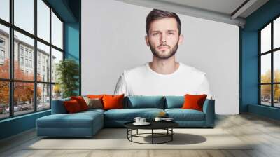 Close up portrait of good-looking serious bearded Caucasian man with blue beautiful eyes wearing white casual t-shirt posing isolated on gray studio wall with copy space for your promotional content. Wall mural