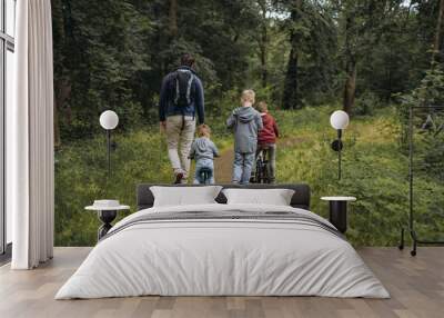 A young caucasian man and three cute boys walking along a path in forest in summer time. One boy is cycling by bicycle, second by runbike, third is riding a kick scooter Wall mural