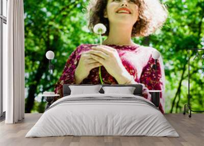 A pretty curly girl with a dandelion in her hands standing in the park Wall mural