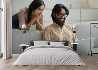 Arabian Indian couple on couch sofa use computer laptop shopping online internet store excited multiracial wife husband happy excited customers woman and man search goods purchase on website at home Wall mural