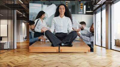 Calm businesswoman female leader woman sitting on table in lotus position meditating in office feels calmness ignore annoyed angry colleagues throw documents business conflict quarrel on background Wall mural